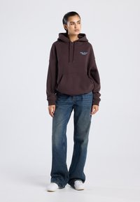 Sudadera marrón con bolsillo frontal y cordón, combinada con jeans azules holgados y zapatillas blancas. Tejido suave, corte relajado, diseño minimalista.