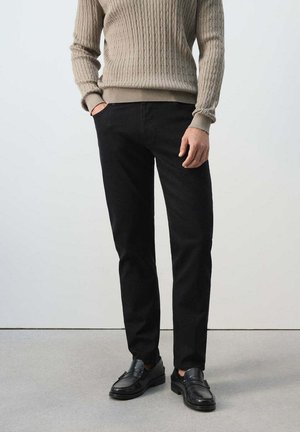 Mann trägt einen beigen Pullover mit Zopfmuster, schwarze gerade Hose und schwarze Loafers und steht mit einer Hand in der Tasche vor einem einfarbigen Hintergrund.