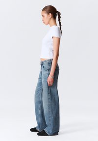 Un crop top blanc à manches courtes associé à un jean taille haute à jambes larges de couleur bleu clair. Le jean présente une taille haute et une finition décolorée et texturée. Des chaussures noires.