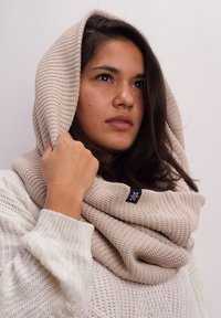 Beige knitted infinity scarf, textured with a ribbed pattern, draped over shoulders, featuring a small label in the corner.