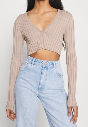 Frau trägt eine beige gerippte, bauchfreie Strickjacke mit Knöpfen und hoch taillierte hellblaue Jeans vor einem schlichten Hintergrund.