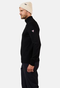 Suéter de punto negro con cuello alto, puños y dobladillo acanalados; presenta un pequeño parche en la manga izquierda. El modelo lleva un gorro crema.