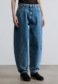 Šviesiai mėlyni, laisvo silueto džinsai iš džinsinio audinio su aukštu liemeniu, penkiomis kišenėmis ir sagų užsegimu, derinami su juodais sportiniais batais.