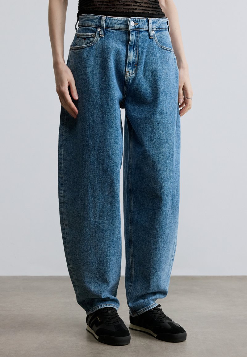 Šviesiai mėlyni, laisvo silueto džinsai iš džinsinio audinio su aukštu liemeniu, penkiomis kišenėmis ir sagų užsegimu, derinami su juodais sportiniais batais.