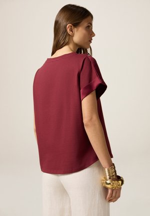 Top corto bordeaux con una vestibilità rilassata, dotato di scollatura rotonda e dettagli in satin ai polsini. Indossato con pantaloni fluidi di colore chiaro.