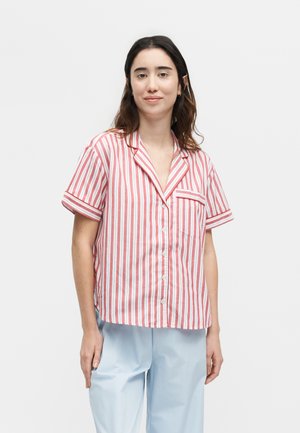 Nő látható egyszínű háttér előtt, vörös-fehér csíkos rövid ujjú, gombos ingben és világoskék nadrágban.