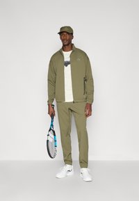 Ελαιόστηθο αθλητική ενδυμασία με ζακέτα με φερμουάρ και παντελόνι που στενεύει, συνδυασμένη με ένα ελαφρύ μπλουζάκι με γραφικά, λευκά αθλητικά παπούτσια και καπέλο.