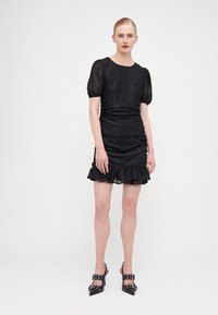 Robe noire texturée avec des manches bouffantes, un corps froncé et un ourlet en volants. Assortie à des escarpins noirs pointus avec des accents de boucle.
