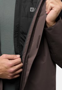 Giacca isolante marrone e nera, con chiusura frontale a zip, morbida fodera interna e chiusura in velcro sulla tasca interna.