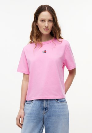 Jeune femme aux cheveux ondulés portant un T-shirt rose uni avec un petit logo et un jean taille haute bleu clair sur fond blanc.