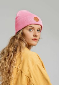 Różowa dzianinowa czapka z ribbowym wzorem i okrągłą brązową skórzaną łatą, noszona z żółtą oversize'ową koszulą, na szarym tle.