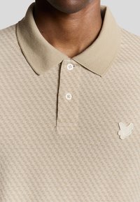 Beige Poloshirt mit strukturiertem Muster, Drei-Knopf-Verschluss und einem Logo-Patch auf der linken Brust. Der Stoff wirkt gestrickt und hat eine glatte Oberfläche.