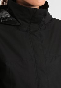 Žena v černé bundě s vysokým límcem, s kapucí vyloženou síťovinou, která je částečně viditelná vzadu.