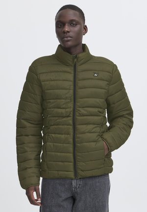 Jeune homme portant une veste matelassée vert olive fermée par une fermeture éclair, une main dans la poche et un jean gris sur un fond uni.