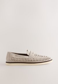 Mocassin homme beige clair avec des détails en cuir tressé sur les côtés et le dessus, doté d’une semelle blanche plate et d’un design sans lacets.