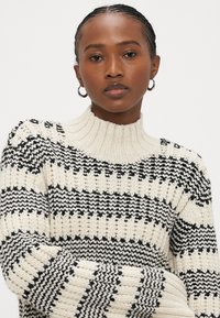 Pletený svetr s vysokým žebrovaným límcem, střídajícími se pruhy v černé a krémové barvě a robustní strukturou látky.