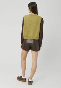 Veste tricotée verte sans manches, associée à un short en cuir marron. Le fond est uni et clair. Chaussures marron avec accents blancs.