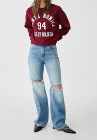 Donna che indossa una felpa bordeaux "Santa Monica 94 California", jeans larghi strappati blu chiaro e scarpe nere con borchie a punta in piedi.