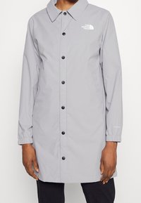 Abrigo ligero gris, hecho de un material resistente al agua. Cuenta con un collar, botones negros y puños elásticos. Logo en el lado izquierdo del pecho.