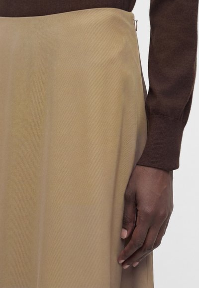 Το δεξί χέρι ακουμπάει δίπλα σε μια μπεζ φούστα, με ένα καφέ μακρυμάνικο τοπ ορατό στο κάδρο.