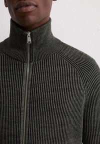 Donkergroen geribde zip-up sweater met een hoge kraag en metalen rits. Het ontwerp heeft een gestructureerd, gestreept patroon door het geheel.