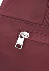 Bolsa de nailon burdeos con un bolsillo con cremallera, que presenta un tirador de cremallera plateado etiquetado con "Bogner" y costuras texturizadas a lo largo de los bordes.
