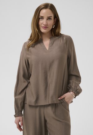 Frau posiert mit einer Hand in der Tasche, trägt eine hellbraune Langarmbluse und passende lockere Hosen vor einem einfarbigen Hintergrund.