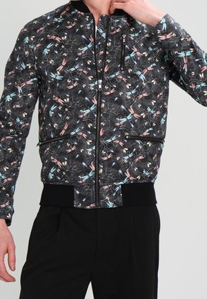 Mężczyzna nosi czarną kurtkę bomber z kolorowym kwiatowym i ważkowym wzorem, czarnymi mankietami i kieszeniami na zamek.