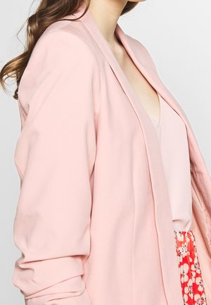 Vrouw die een lichtroze blazer draagt over een lichtroze top en een rode bloemenrok, met lang bruin golvend haar zichtbaar tegen een effen achtergrond.