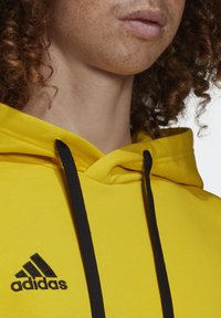 Gros plan d'une personne portant un sweat à capuche jaune Adidas avec des cordons noirs, montrant les lèvres, le cou et les cheveux bouclés.