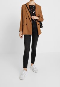 Brun dubbelknäppt blazer över en svart topp med ett färgglatt mönster, kombinerad med svarta skinny jeans och vita sneakers.