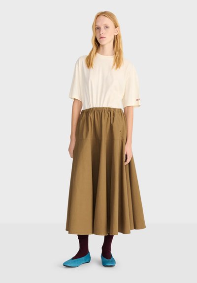 Jeune femme aux longs cheveux blonds porte un haut crème ample, une jupe marron taille haute, des chaussettes bordeaux et des chaussures plates bleues, debout sur un fond uni.