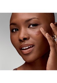 Zbliżenie na osobę o gładkiej, ciemnobrązowej skórze, która nakłada produkt do pielęgnacji skóry na policzek. Ma krótkie włosy i neutralny lakier do paznokci.