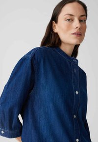 Camicia in denim blu scuro con colletto, maniche lunghe arrotolate fino ai gomiti e bottoni a pressione perlescenti lungo la parte anteriore. Tessuto liscio.