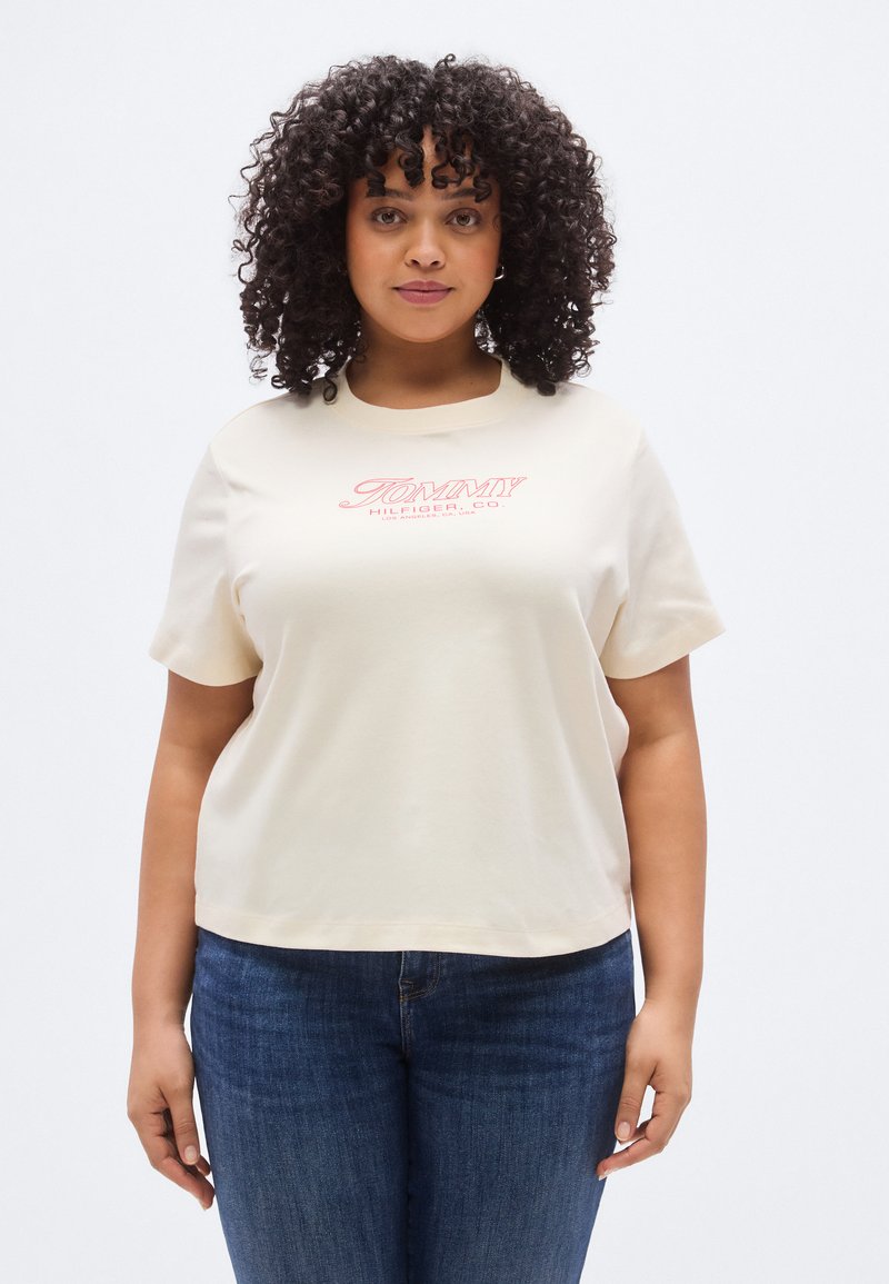 Μπεζ κοντομάνικο μπλουζάκι από βαμβάκι, με στρογγυλή λαιμόκοψη και μπροστινό λογότυπο σε κόκκινο κείμενο. Φορεμένο με παντελόνι από σκούρο τζιν.