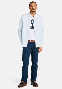 Uomo in piedi, vestito con una camicia azzurro chiaro sopra una maglietta bianca con grafica, pantaloni blu navy, cintura marrone e scarpe marroni scure su sfondo bianco.