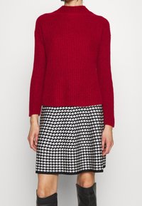 Suéter rojo de canalé con un cuello alto, combinado con una falda de toile de Jouy en blanco y negro. El atuendo presenta mangas largas y una silueta ajustada.