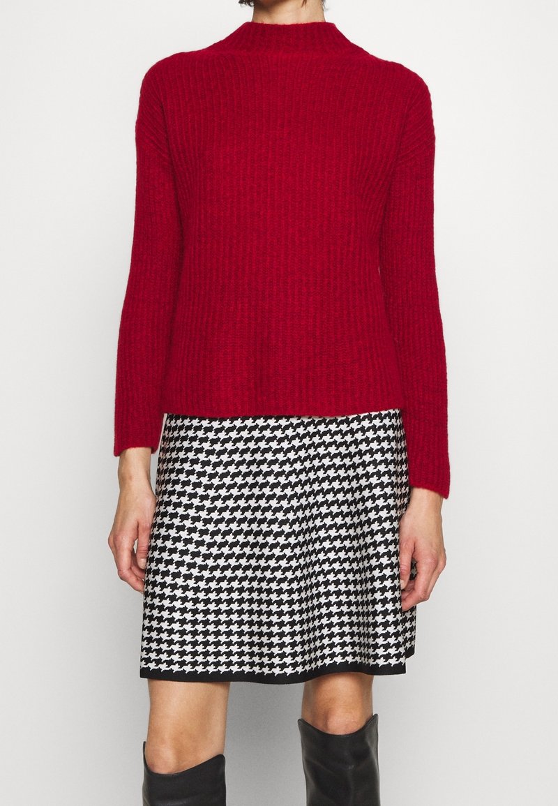 Suéter rojo de canalé con un cuello alto, combinado con una falda de toile de Jouy en blanco y negro. El atuendo presenta mangas largas y una silueta ajustada.