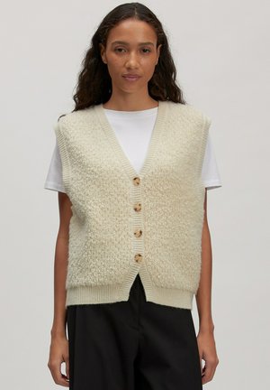 Vrouw in een crèmekleurige textuur gelaagde vest met knopen over een witte korte(mouwen) shirt en zwarte broek, staand tegen een effen achtergrond.