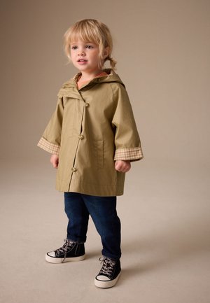 Niño pequeño con el pelo rubio de pie, vestido con un abrigo beige con capucha y puños a cuadros, vaqueros oscuros y zapatillas negras altas con estampado.