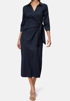 Kobieta w granatowej sukience midi o kroju kopertowym z trzy-czwarte rękawami i wiązaniem z boku, w połączeniu z beżowymi czółenkami z paskami.