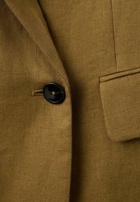 Gros plan sur une veste en tissu marron texturé avec un bouton noir et un rabat de poche rectangulaire.