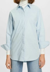 Ljusblå button-up skjorta i bomull, med långa ärmar och uppvikta manschetter samt en klassisk krage, stylad över en vit höghalsskjorta.