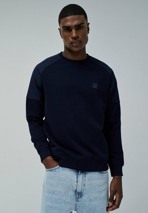 Sudadera navy con cuello redondo acanalado, mangas raglán y un pequeño parche con el logo. Combinada con pantalones de mezclilla azul claro. Textura suave.