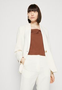Bílý blazer se zlatým knoflíkem, hnědý saténový top a bílé upravené kalhoty. Hladké textury; uvolněný střih, minimalistický design. Neformální, ale přesto elegantní.