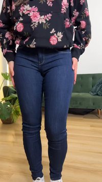 Personne portant un jean skinny bleu foncé et un haut à manches longues noir à motif floral, debout à l'intérieur près d'un canapé vert et de plantes d'intérieur.