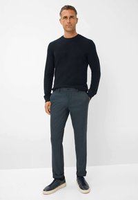 Pullover a coste blu navy, pantaloni grigi scuri su misura e sneaker scure. I pantaloni hanno un dettaglio con bottone; le textures sono lisce e aderenti.