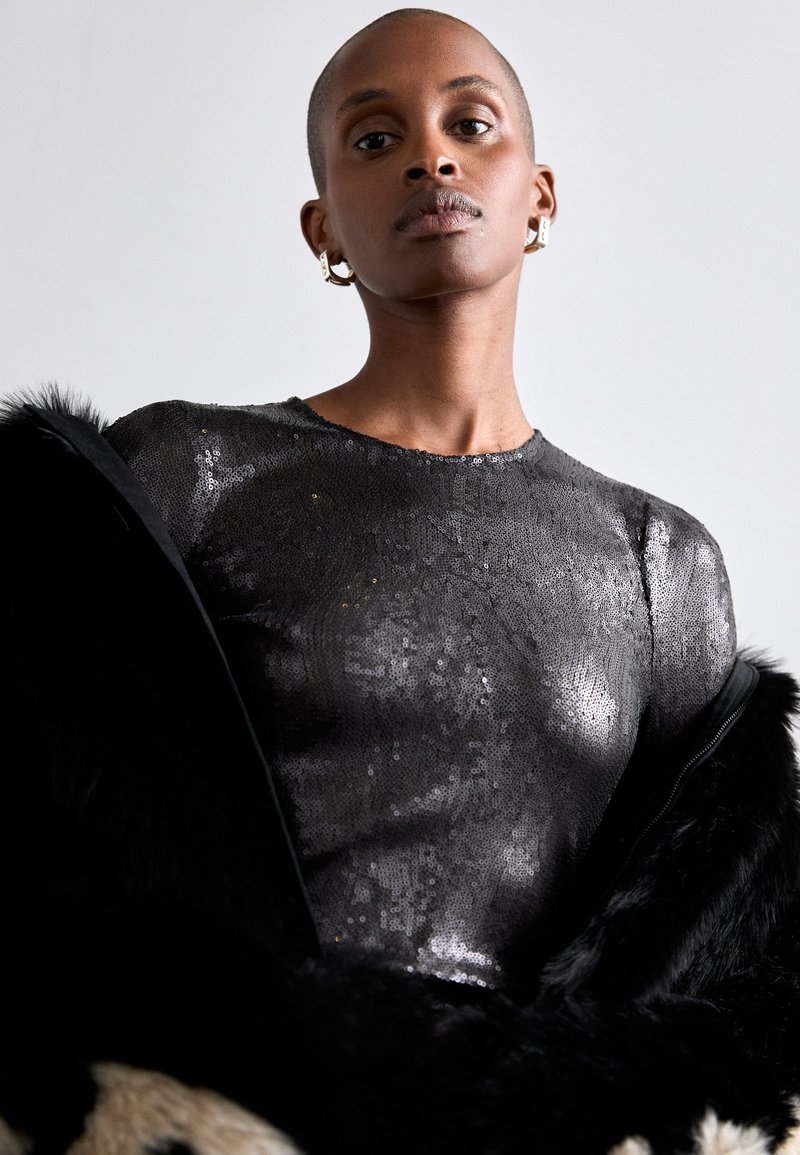 Haut à sequins noirs avec un col rond, à coupe ajustée. Le modèle porte un manteau en fourrure noir, mettant en avant une texture brillante et lustrée.