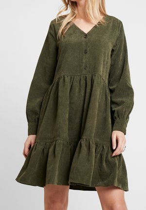 Frau trägt ein olivgrünes Cordkleid mit langen Ärmeln, Knöpfen, geraffter Taille und Rüschensaum.