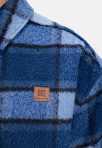 Blue plaid flannel shirt featuring a soft, fuzzy texture, contrasting dark lines, and a brown leather label on the chest.
